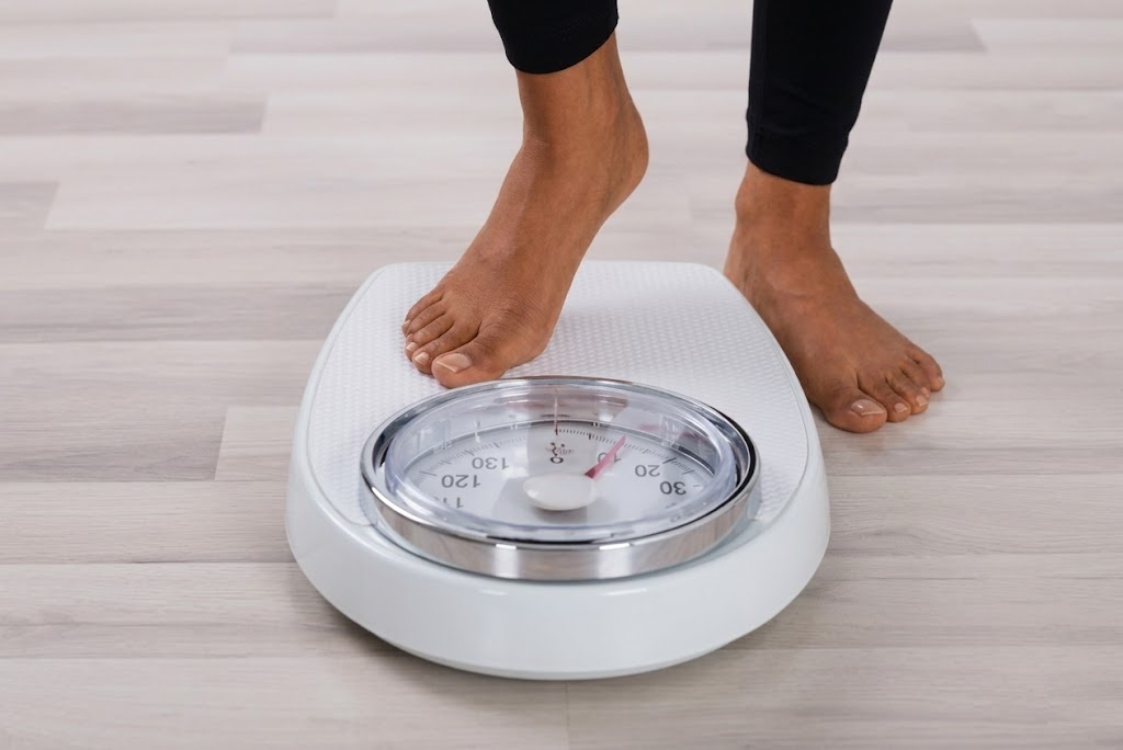 A low-angle close-up of a person's dark-skinned feet and legs in black leggings as they step onto a white mechanical bathroom scale on a light wood floor, with the analog dial visible in the foreground.